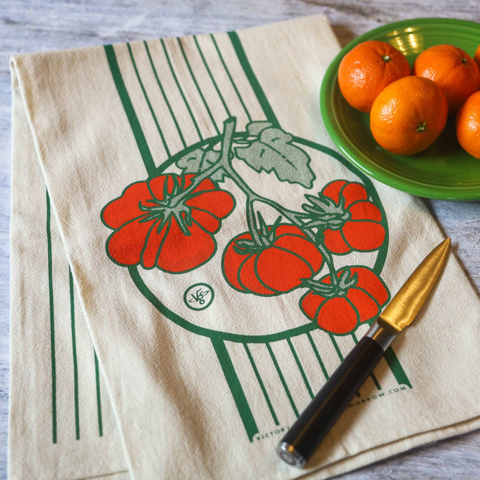 Beige towel with tomato design, knife, and oranges on a wooden surface